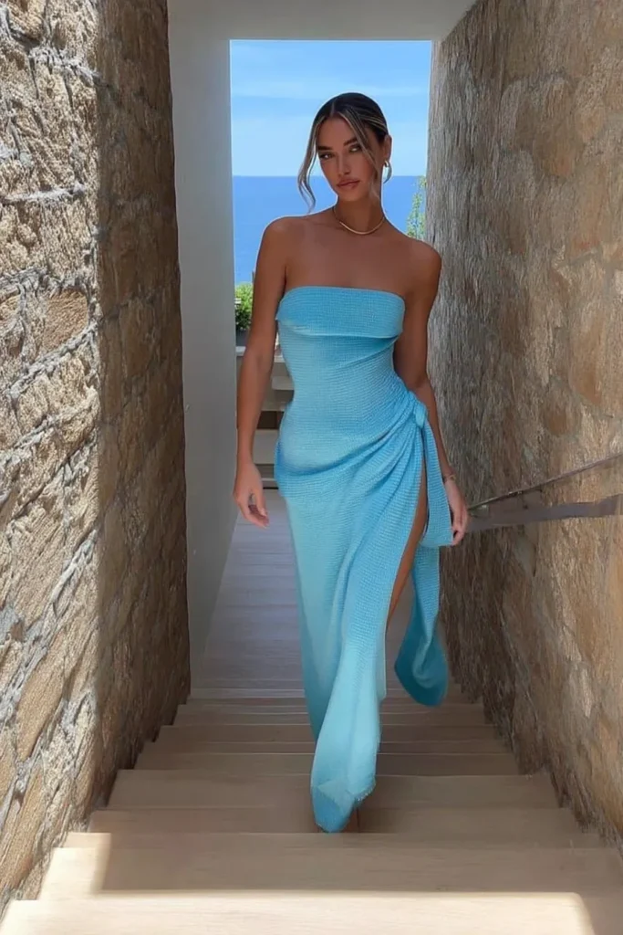 Elegant pastel blue strapless bandeau maxi dress with side slit and gold necklace perfect for glamorous island vacation outfits in Mediterranean stone corridor