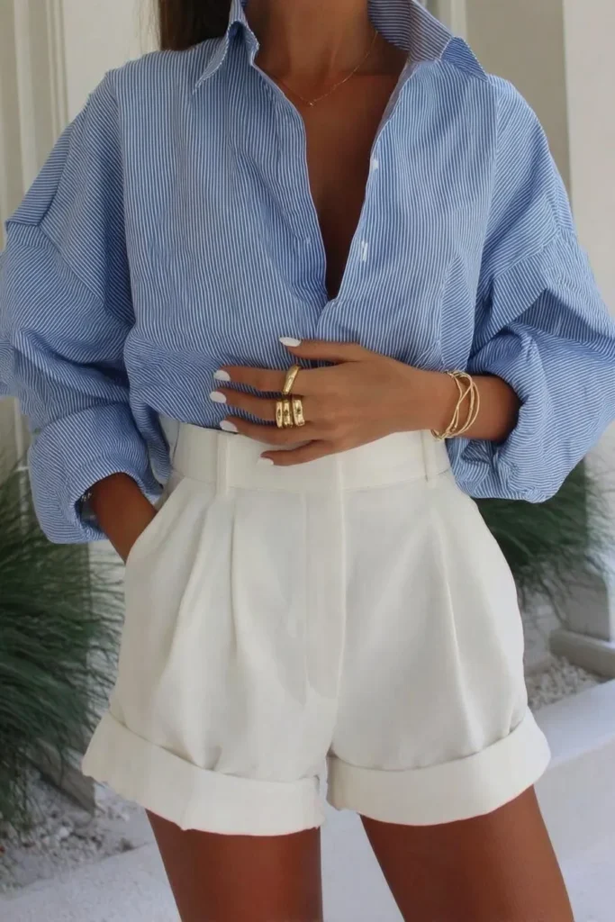 Chic blue casual spring outfits displaying light blue and white striped collared shirt with shorts and white leather belt, complete with gold rings for timeless minimalist style