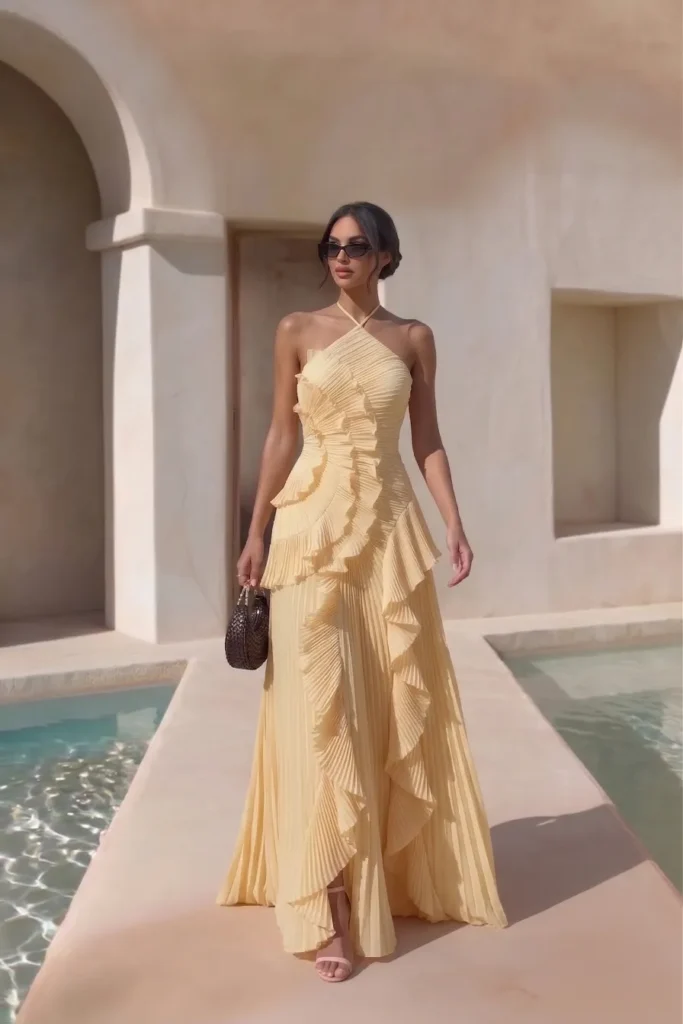 Chic butter yellow trendy wedding guest dress, halter neckline, cascading ruffle panels and pleated maxi skirt, accessorized with a handbag, sunglasses  for  luxury  wedding look