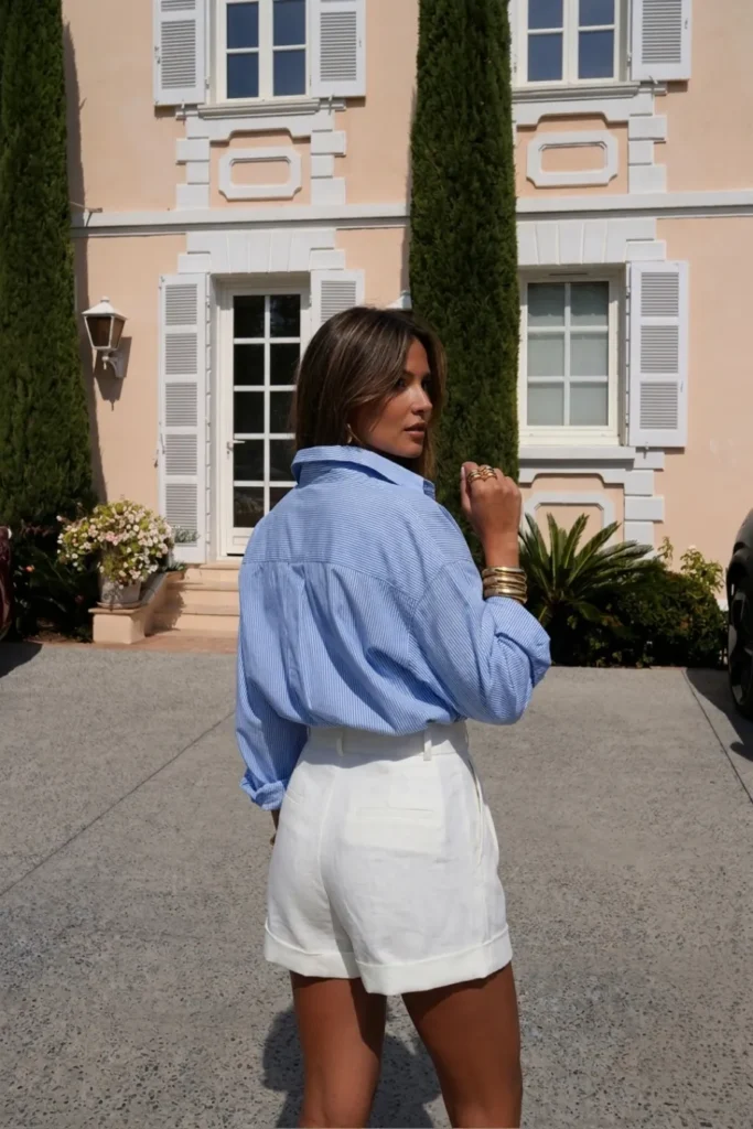 Chic blue outfit inspo summer style with an oversized blue striped button-down shirt tucked into tailored white belted shorts,  with gold jewellery, white  sunglasses and strappy white heels