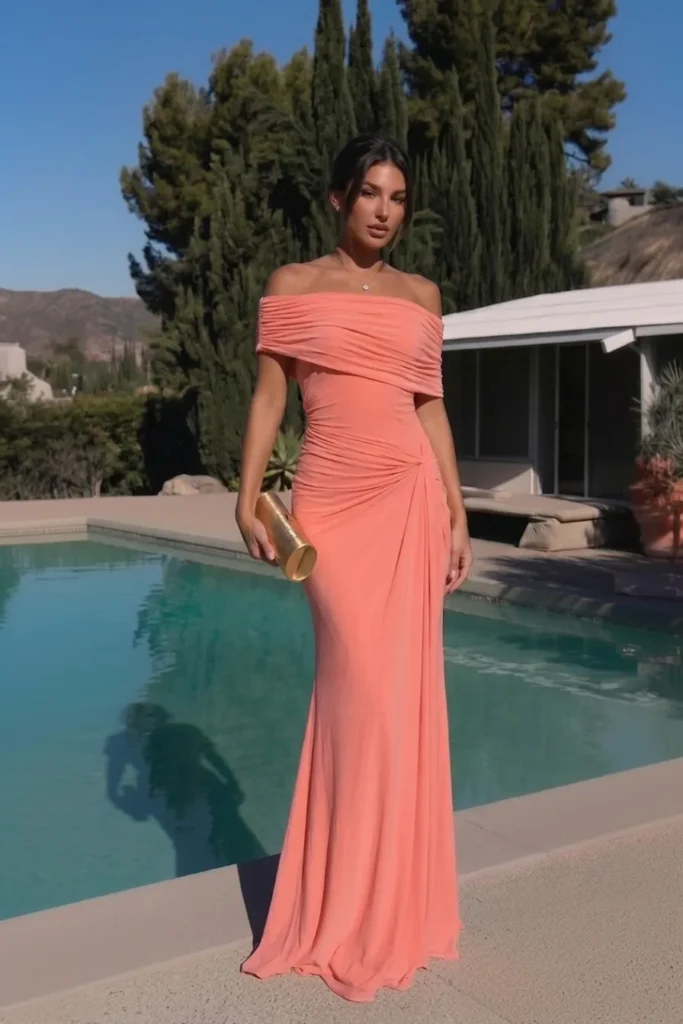 Coral elegant wedding guest dress with an off-the-shoulder ruched neckline, knot waist detail and a fitted floor-length skirt, accessorised with stacked gold bangles and a gold clutch bag
