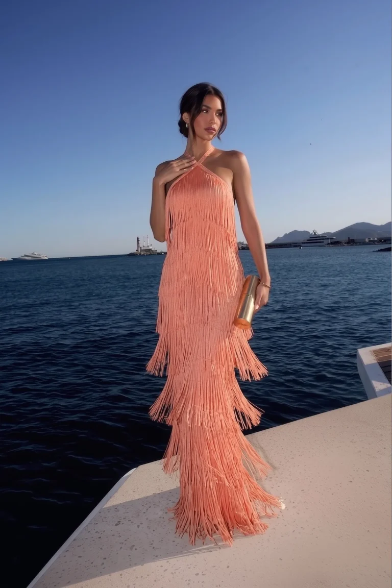 Coral trendy wedding guest dress with a crossover halter neckline and tiered floor-length fringe design, cascading layers of tassels from bodice to hem, accessorised with a gold bag