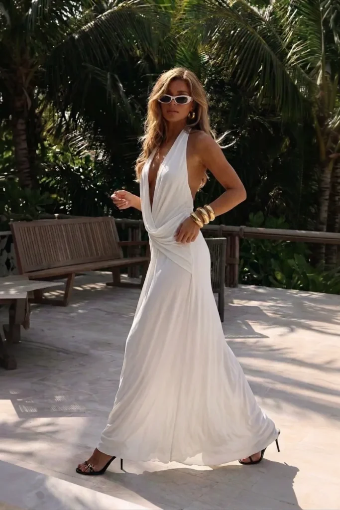 Elegant white summer night outfit, halter neck maxi dress with a draped twisted waist detail, flowing full-length skirt, with gold bangles, white cat-eye sunglasses and black strappy heels